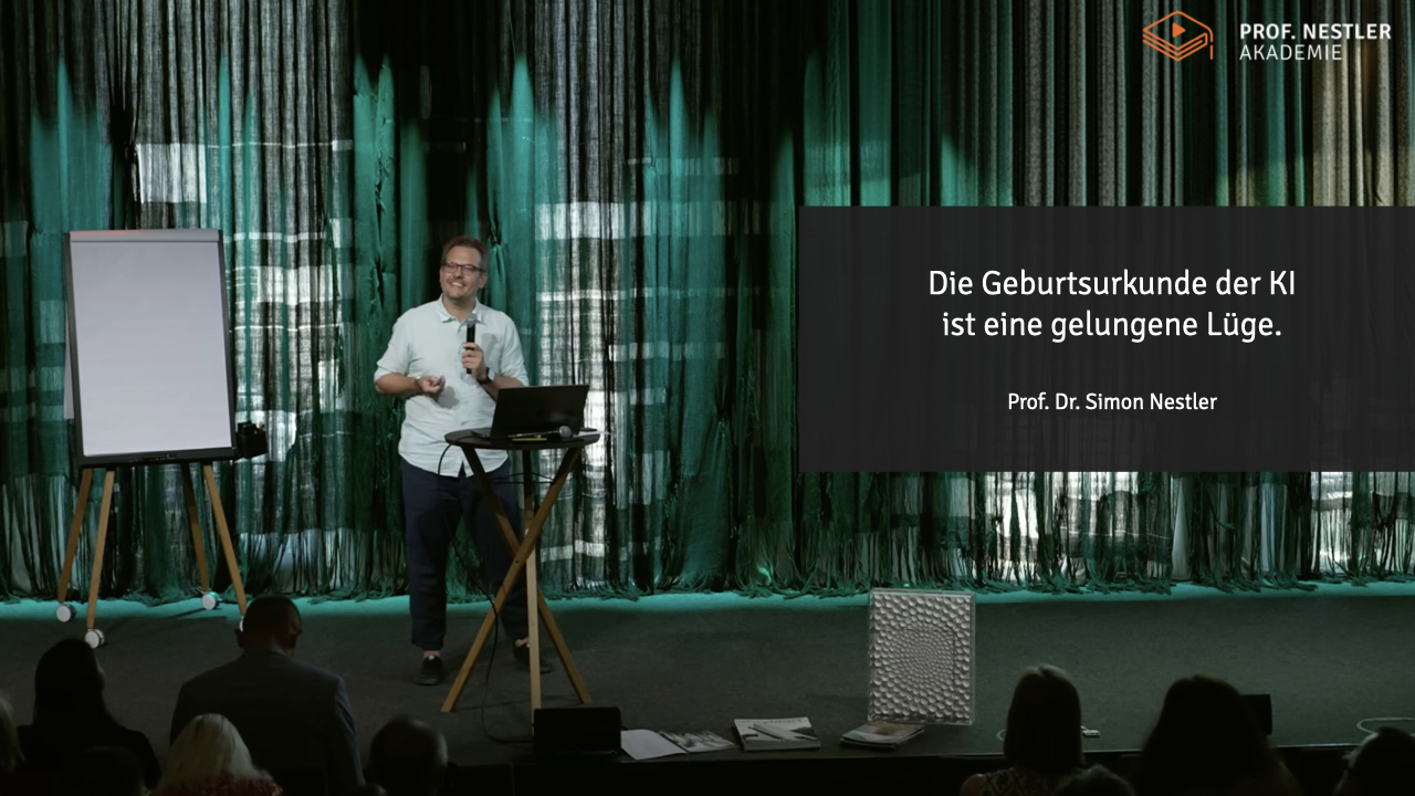 Simon Nestler steht mit Handmikrofon auf einer Bühne vor einem grünen Vorhang, neben ihm ein Stehtisch mit Laptop und links ein leeres Flipchart. Rechts auf der Leinwand die Titelfolie: „Die Geburtsurkunde der KI ist eine gelungene Lüge." Im Vordergrund die Hinterköpfe der ersten Zuschauerreihe.