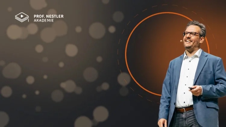 Simon Nestler steht lächelnd auf einer Bühne, im blauen Sakko, mit Headset-Mikrofon und Presenter in der Hand. Hinter ihm ein dunkler Hintergrund mit unscharfen Lichtkreisen und einem orangen Ring. Oben links das Logo der Prof. Nestler Akademie.